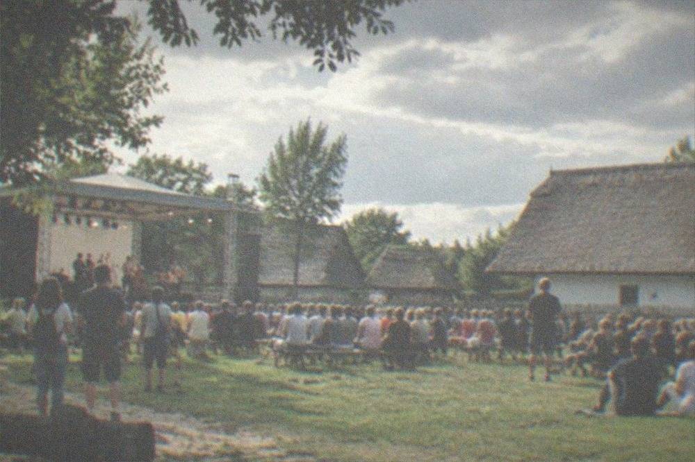 An open-air festival stage and crowd on a museum lawn with traditional houses in the background