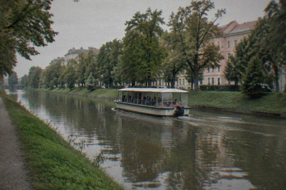 A small passenger water bus moving along a canal between green banks in a European city