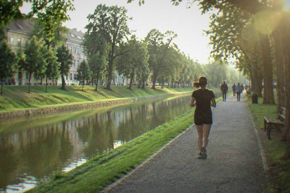 People walking a paved riverside path beside a canal with lawns and old buildings beyond