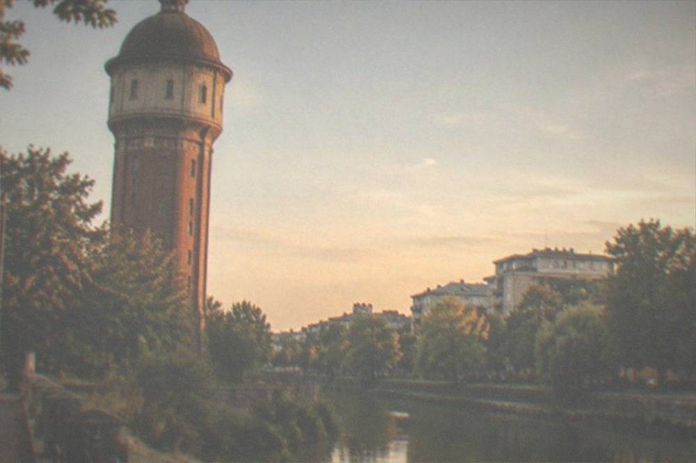 An old stone water tower beside a canal with apartment buildings and trees in the background