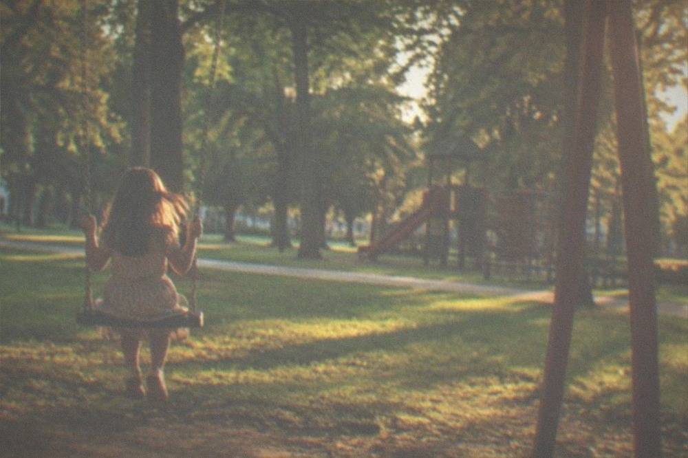 A child on a swing beneath large shade trees in a city park