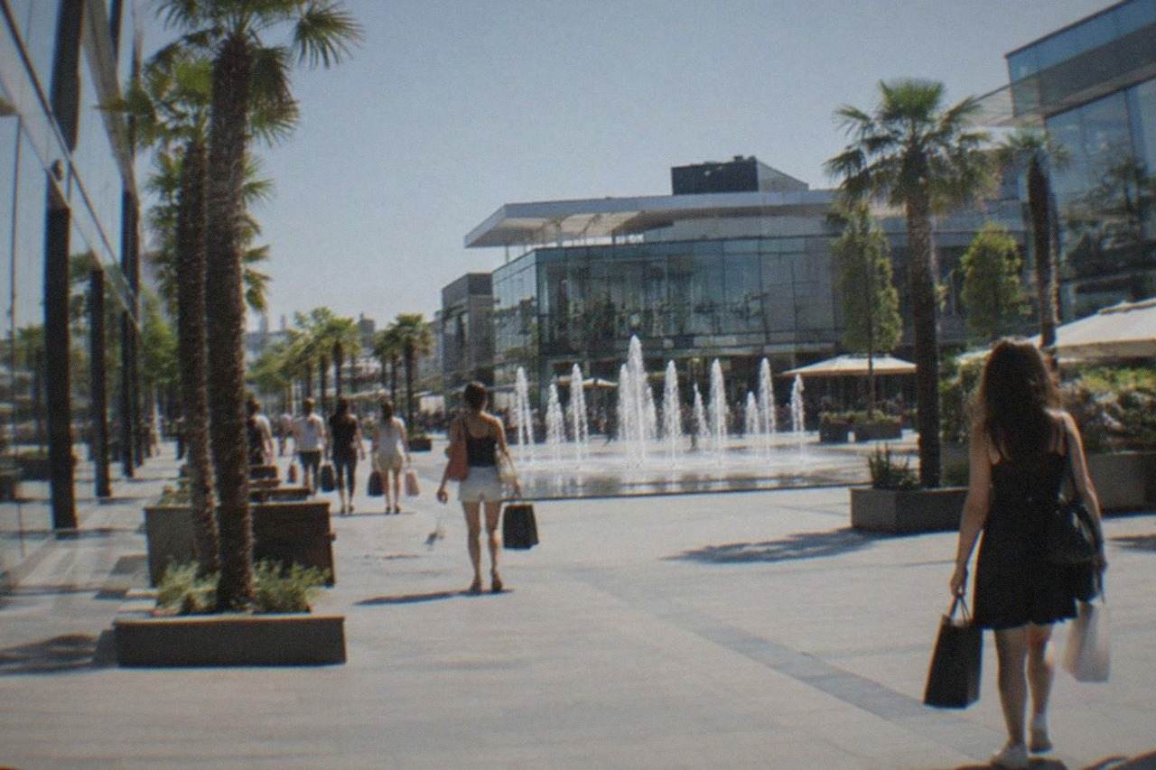 Modern glass shopping mall exterior with palm-lined walkway and people carrying shopping bags