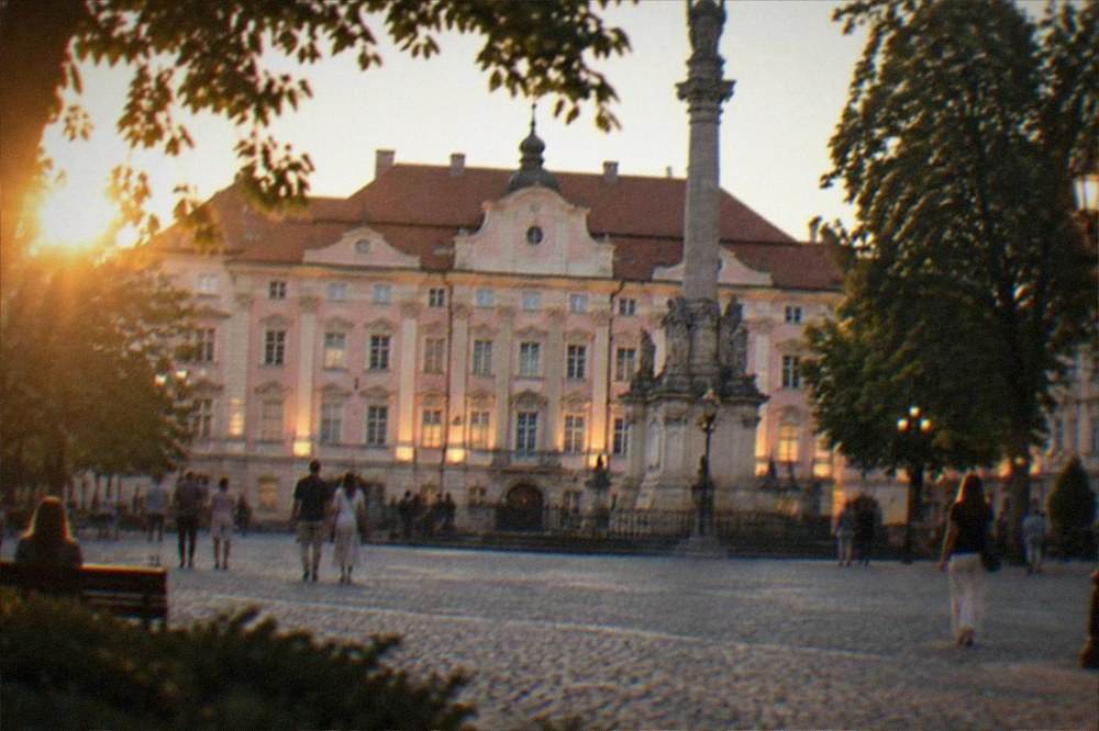 An evening city square with lit facades and people strolling under trees