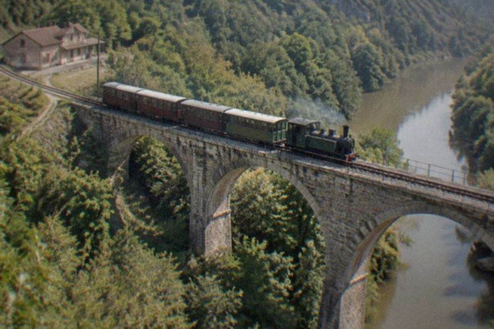 A narrow-gauge train crossing a tall stone viaduct above a wooded valley