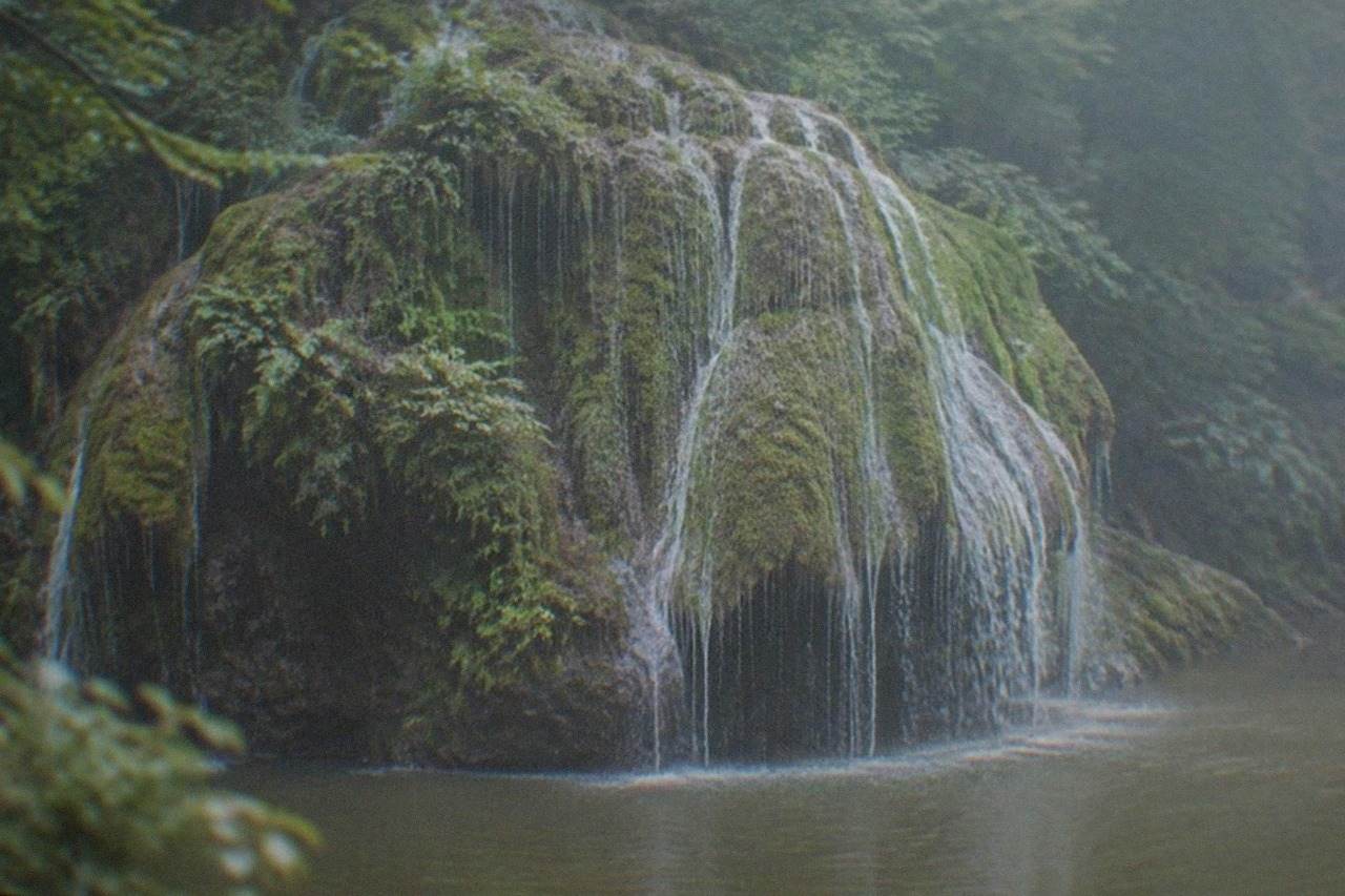 A curtain of water spreading evenly over a moss covered rock dome and falling into a green forest pool