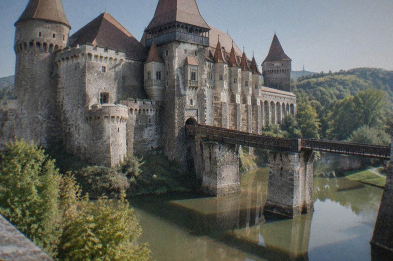 A Gothic castle with tall towers and stone bridges reflected in a moat, seen from the shore on a clear day