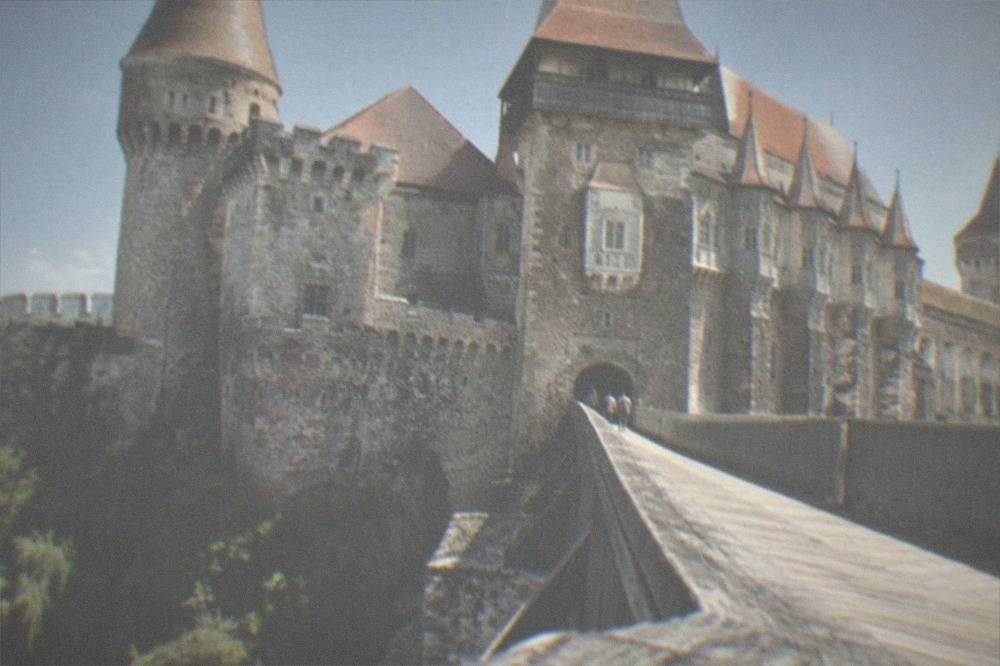 A long wooden footbridge crossing water toward a castle gate with tall stone towers