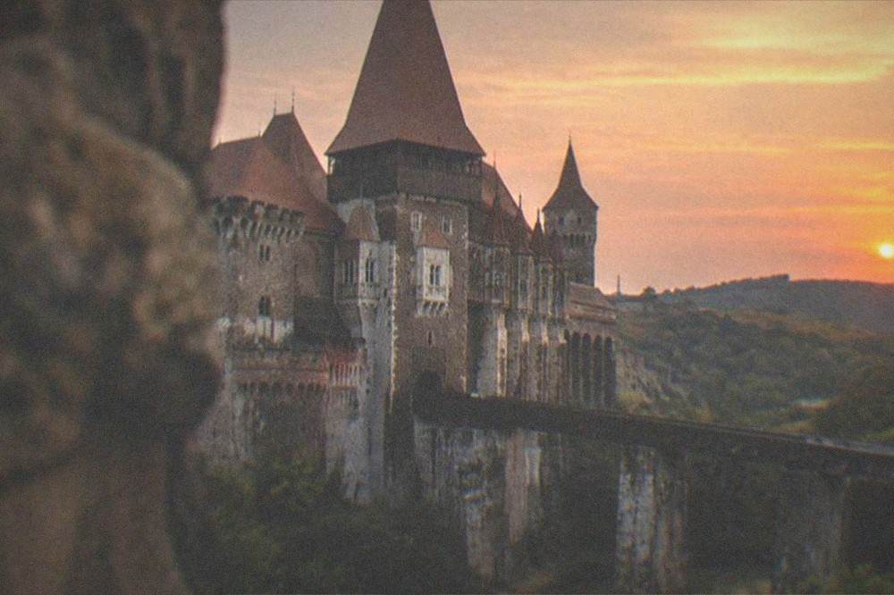 A castle silhouette with pointed towers against an orange sunset sky above dark hills