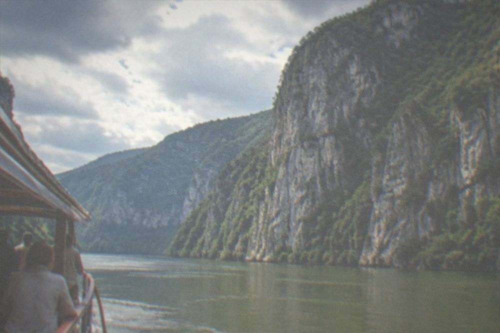 A wooden sightseeing boat on wide green water with tall grey cliffs behind it and passengers at the rail