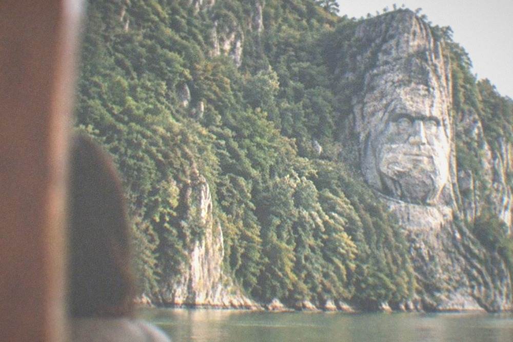 A giant stone face carved into a high cliff beside a wide river with forest on the far bank