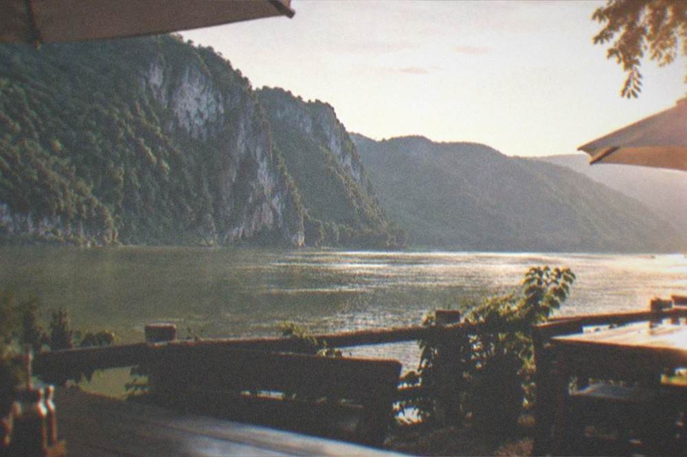 A riverside terrace with simple wooden tables, umbrellas and a view of cliffs across the water