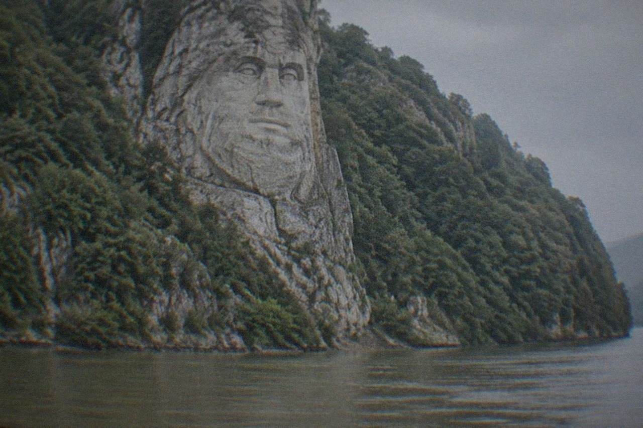 A colossal carved face of a bearded king looking out over the wide Danube river from a rocky cliff