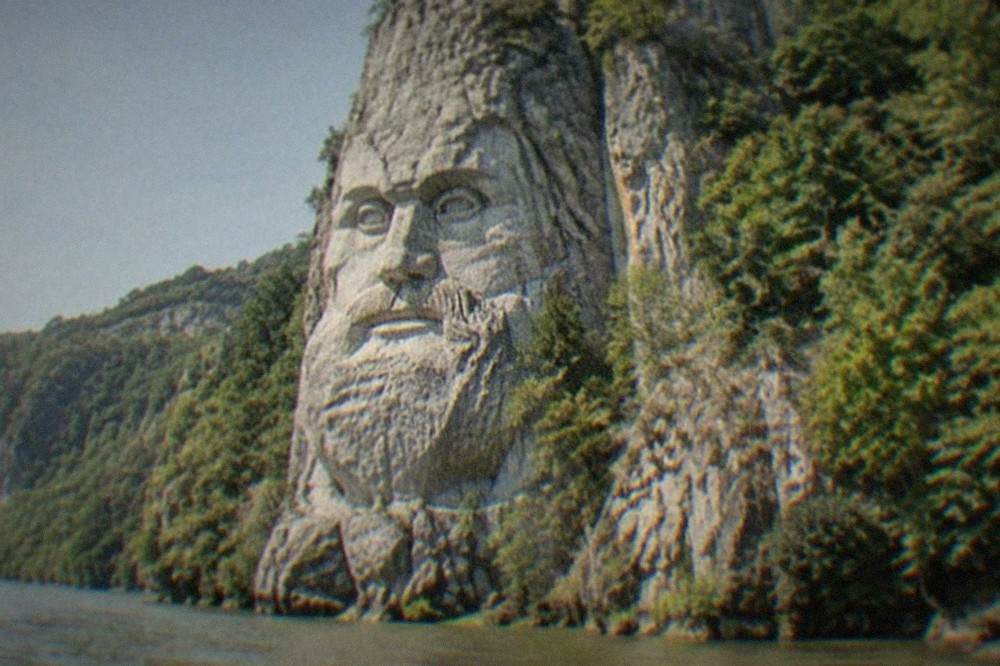 A close view of the carved stone face of a bearded ancient king on a cliff wall above the water