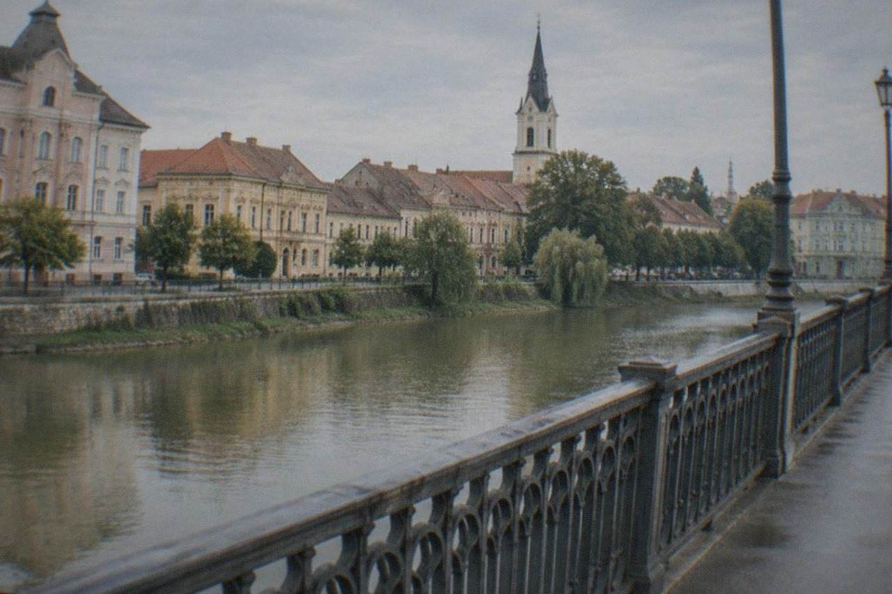 A small Banat town seen from a river bridge, with old buildings and a church tower above the rooftops