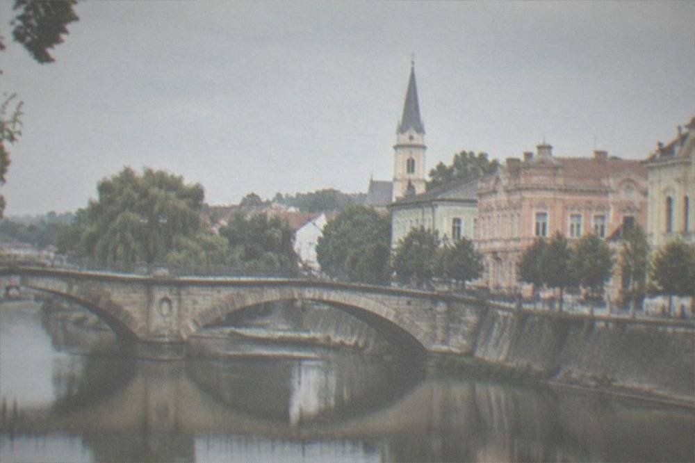 An old stone bridge over a calm river, with trees along the bank and reflected facades in the water