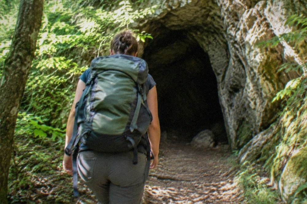 A hiker with a backpack entering a cave entrance framed by green forest