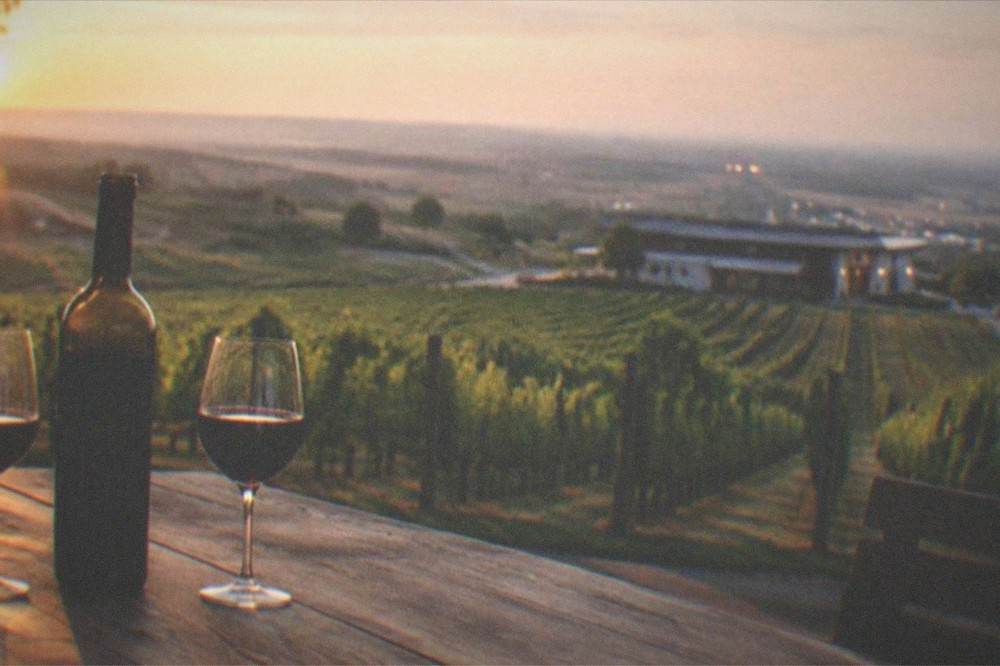 A bottle of red wine and two filled glasses on a wooden table at dusk, with city lights blurred behind