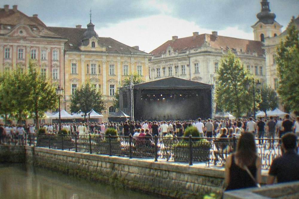A busy festival crowd in a Timisoara square with a stage and historic buildings behind