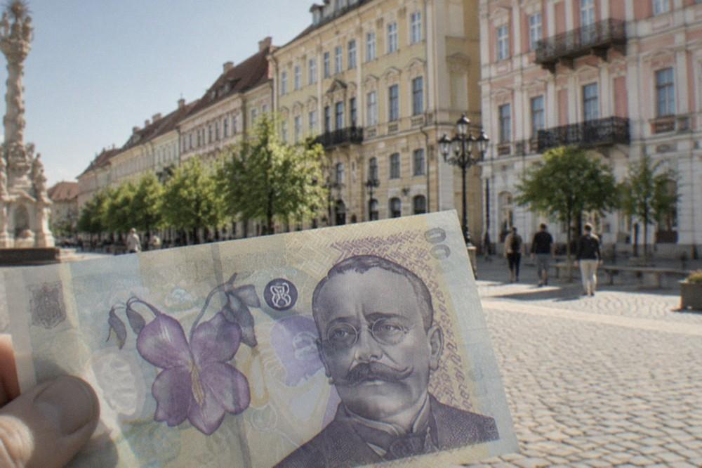 Close-up of a Romanian 100 lei banknote held between fingers in front of a pastel building facade