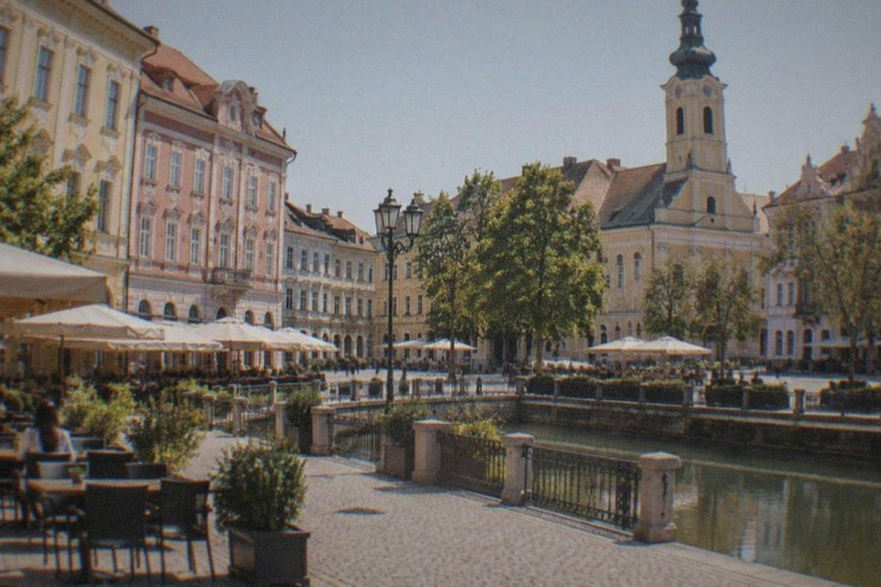 Pastel-colored baroque buildings around a city square with cafe tables and a church tower in the background