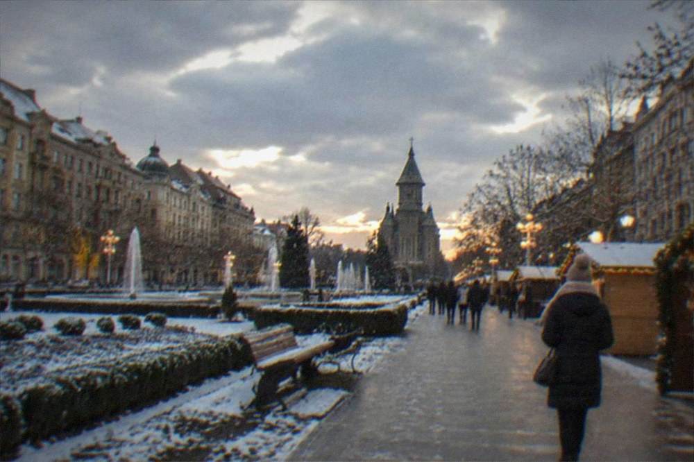 A person in a winter coat and scarf walking past lit holiday stalls in the evening
