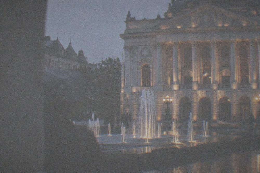 A city square at dusk with an illuminated opera house facade and lit fountains